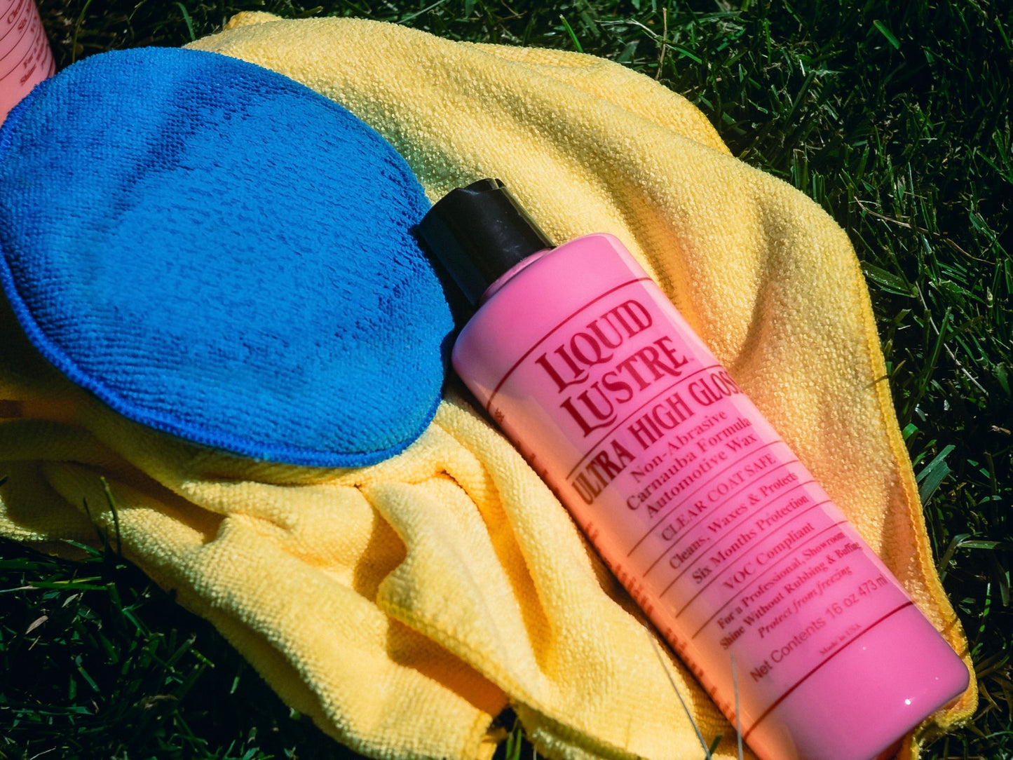 A bottle of The Liquid Lustre Original Pink Wax on a yellow towel with a blue pad, on grass.