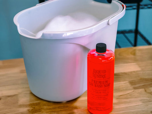White bucket with a bottle of The Liquid Luster car wash soap on a wooden surface.